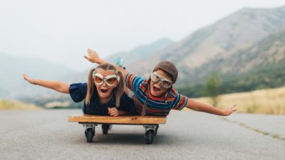 Two kids wearing pilot's goggles take flight down the runway on their tummies with arms outstretched like wings.