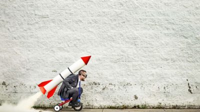 Man with a rocket strapped to his back on a tiny bicycle ready to take off.