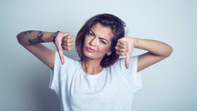 ASX shares downgrade A young woman with tattoos puts both thumbs down and scrunches her face with the bad news.