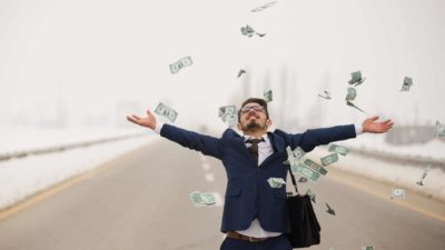 A businessman on a road raises his arms as dollar notes rain down on him.