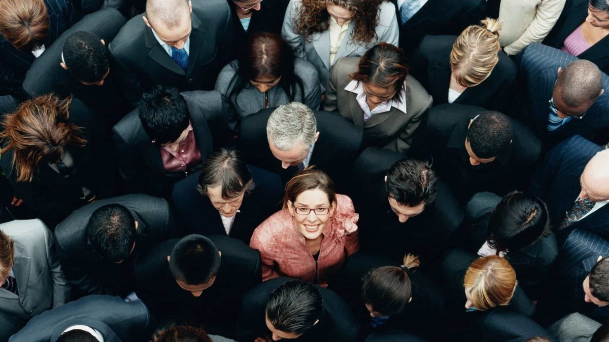 A woman in a crowd of executives stands out as she looks up and smiles.