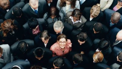 A woman in a crowd of executives stands out as she looks up and smiles.