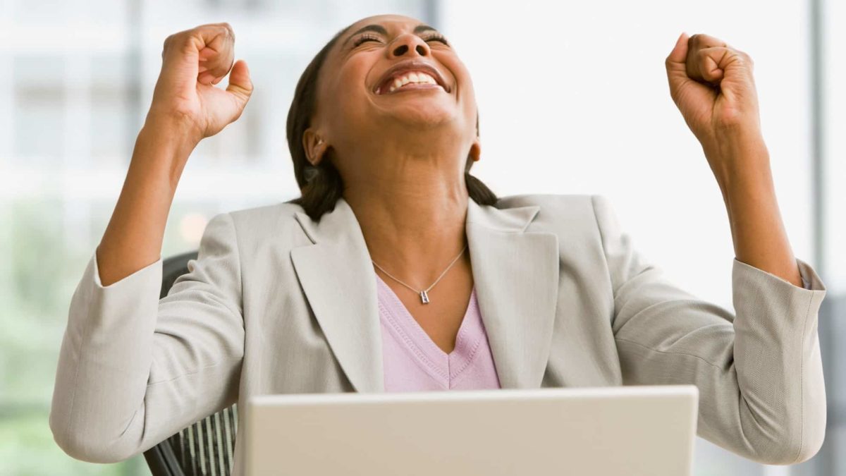 A woman puts heads back and fists in the air as she cheers at laptop.
