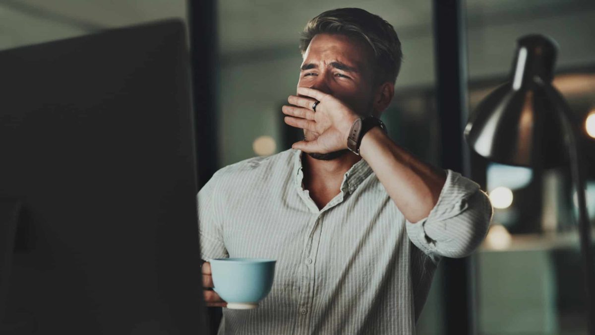 a man sits at his computer screen with a cup of coffee in one hand and the other shielding the bottom half of his face with his eyes closed although he is recoiling from bad news.