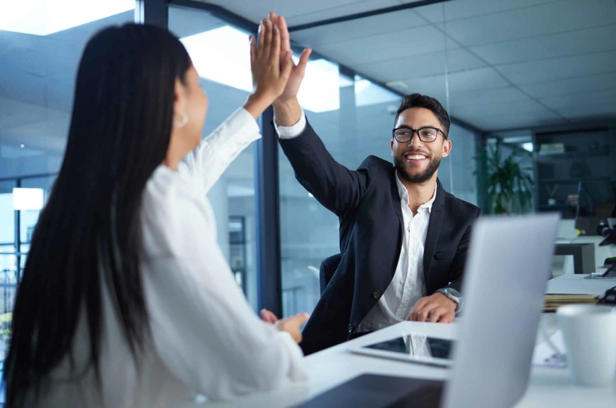 two colleagues high five each other as they sit side by side at a long desk in front of their laptop computers in an office environment.