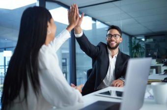 two colleagues high five each other as they sit side by side at a long desk in front of their laptop computers in an office environment.
