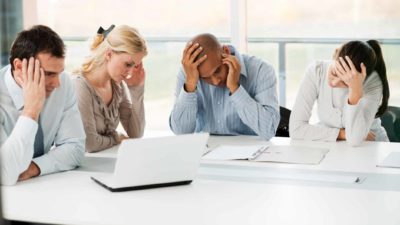 A group of four people, coworkers, sit together looking dejected around a desk with a computer on it in an office setting.