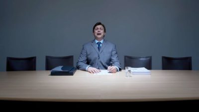 a business person sits at a boardroom table with a sad and apologetic look on his face with empty chairs around him and papers on the desk in front of him.