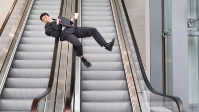 A man in a business suit slides down the handrails of a bank of steel escalators, clutching his documents and telephone.