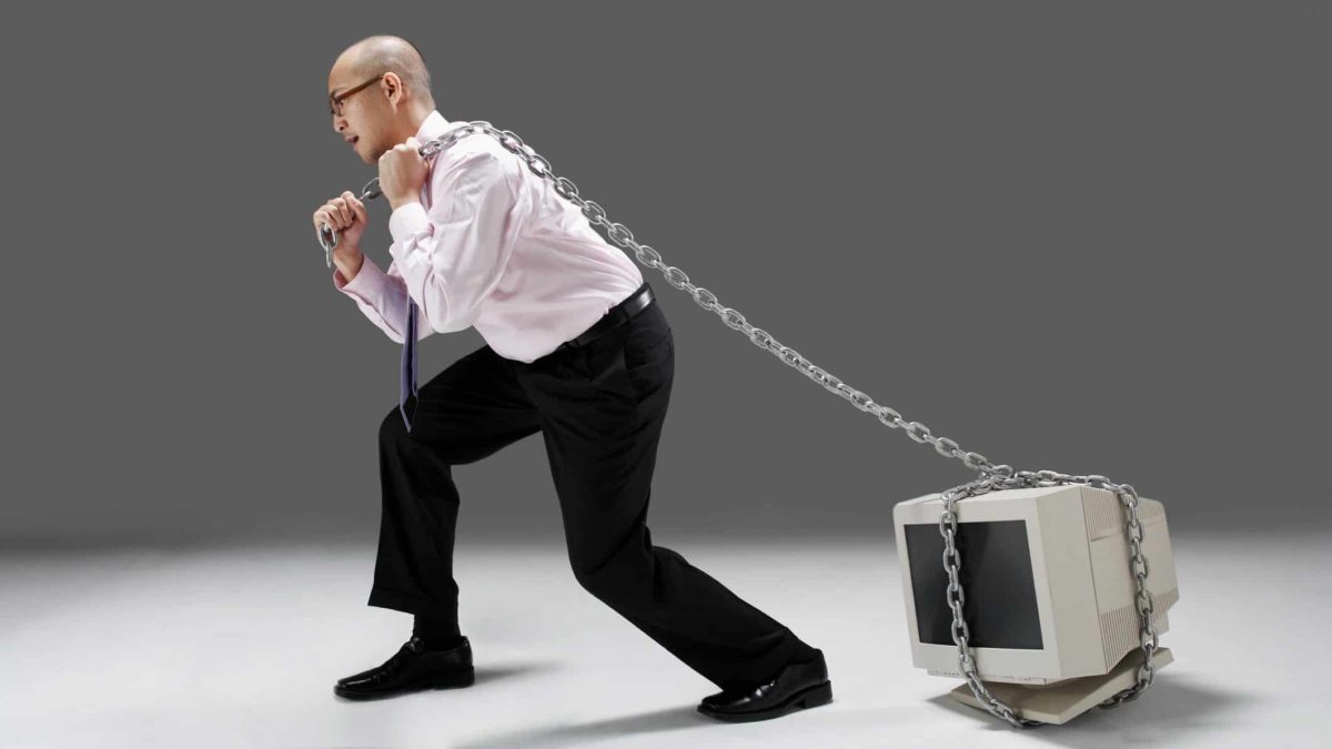 A man in a business shirt and trousers drags a chain wrapped around a computer as thought it is very heavy to move.