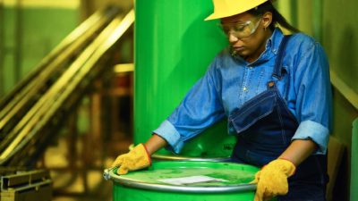 an oil worker with hard hat manoeuvres an oil barrel in an oil production facility.