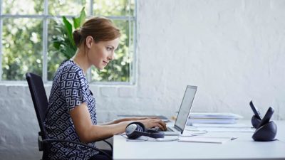 a woman sits at a computer with a satisfied expression on her face in a white room with greenery outside her window.