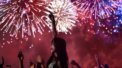 a young woman raises her arm in celebration against a backdrop of brightly coloured fireworks in the sky.