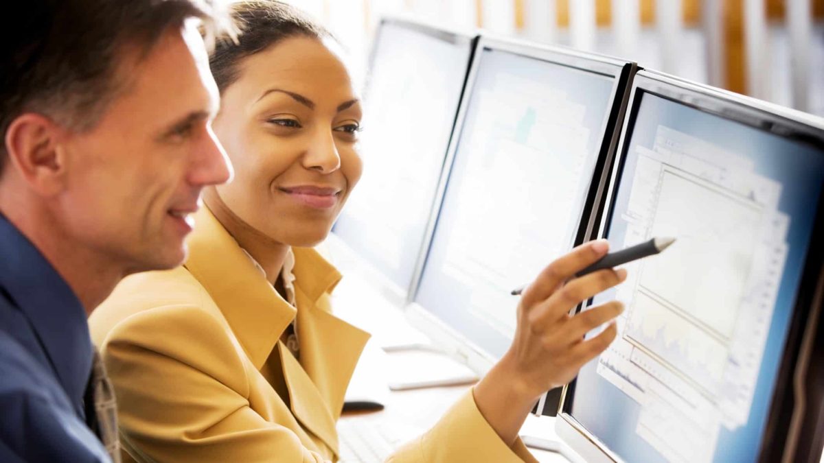 A woman points with her pen at a computer where a colleague sits as though they are collaborating on a project. She has a smile on her face.