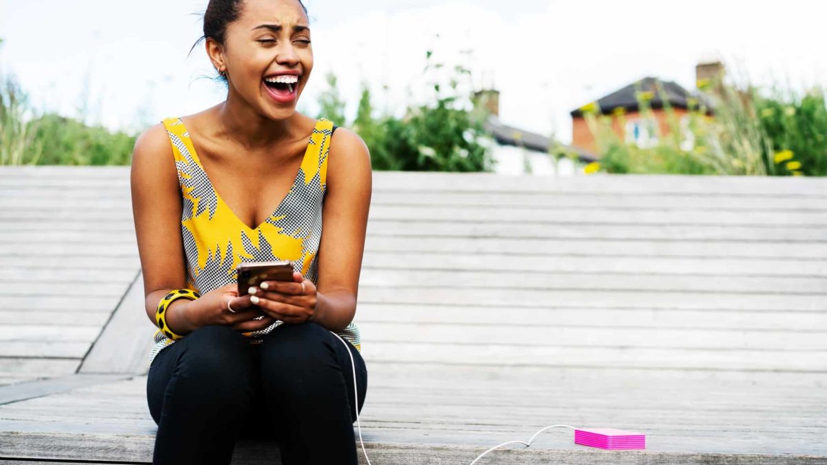 A woman sits on a step laughing at something on her mobile phone as it is being charged by a lithium-powered battery.