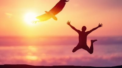 The sunset silhouette of a person leaping in the air as a large bird flies over head.