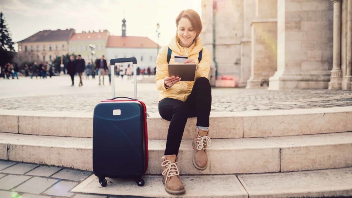 A young woman makes an online travel booking as she sits on some steps with her suitcase next to her.