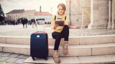 A young woman makes an online travel booking as she sits on some steps with her suitcase next to her.