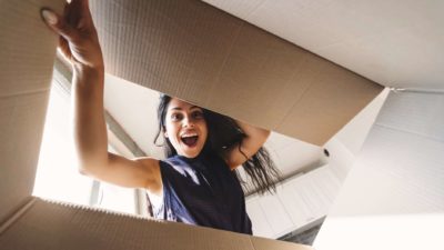 A woman opens and reaches into a big cardboard box, with a big smile on her face.
