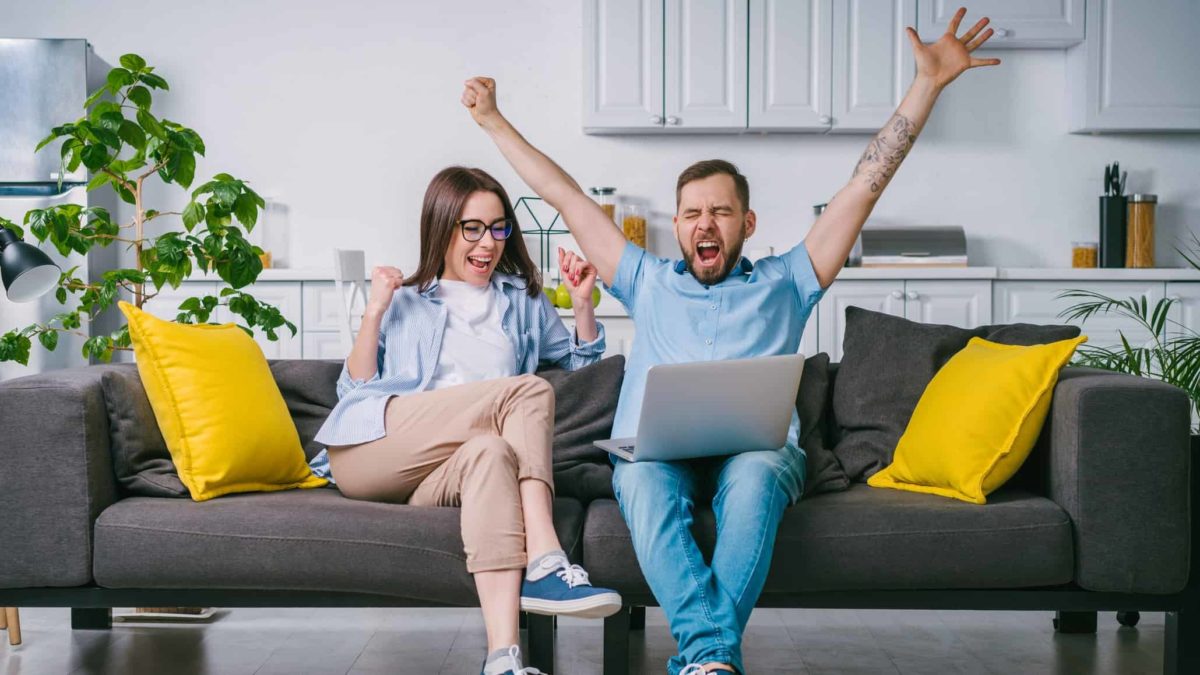 A couple cheers as they sit on their lounge looking at their laptop and reading about the rising Redbubble share price