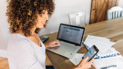 A smiling woman compares broker fees on her laptop and mobile phone.