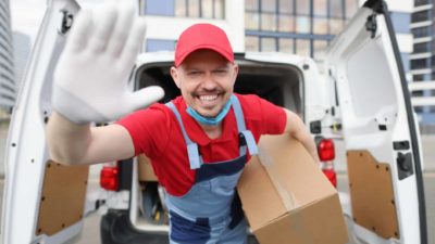 Happy courier driver smiles and waves with a white glove on his hand as he holds a box for delivery with the back of his van in the background.