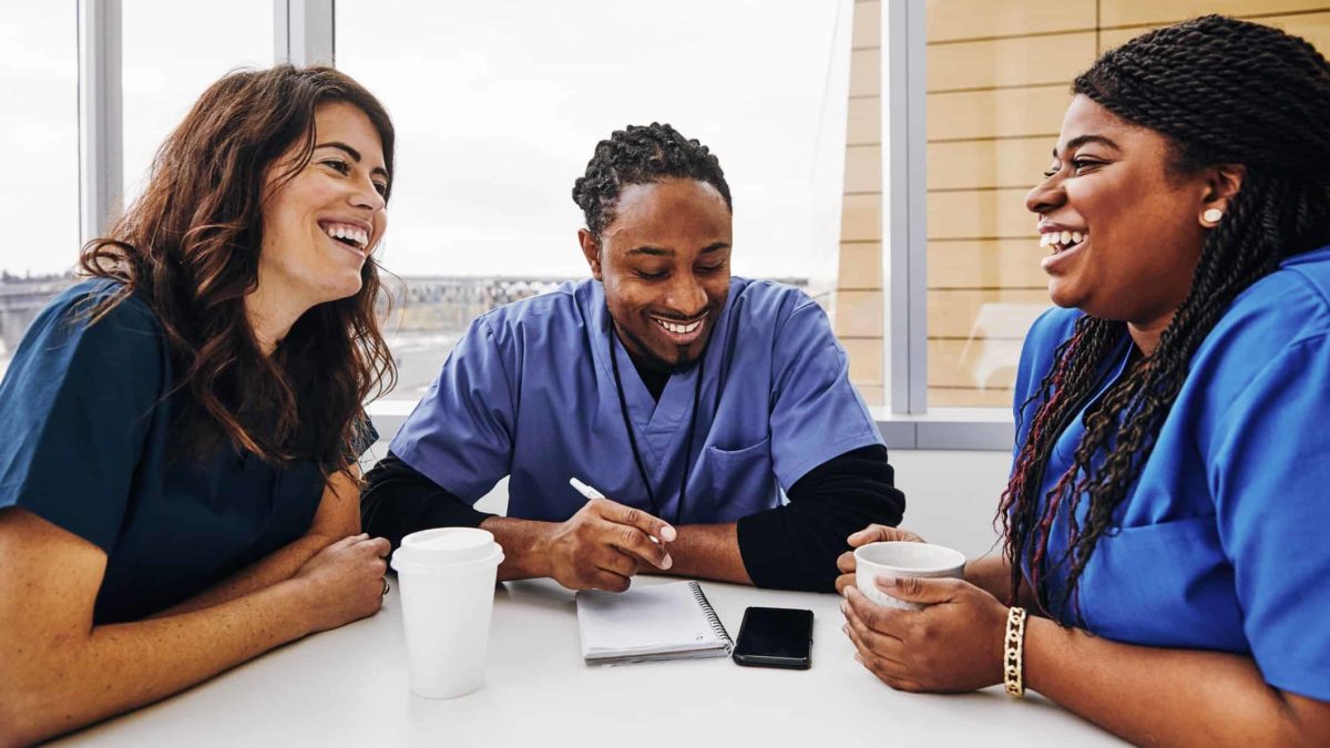 Three medical staffers sit at a table and chat happily wearing hospital scrubs