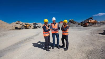 Three men on a mining site wearing hard hats discussing plans