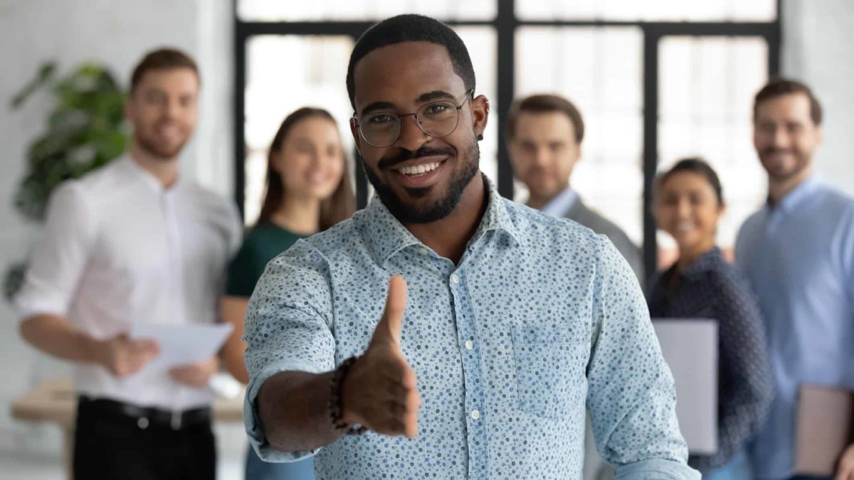 A man standing in front of co-workers extends his hand in welcome