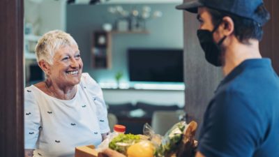 A delivery driver hands over groceries to an older woman at her front door.