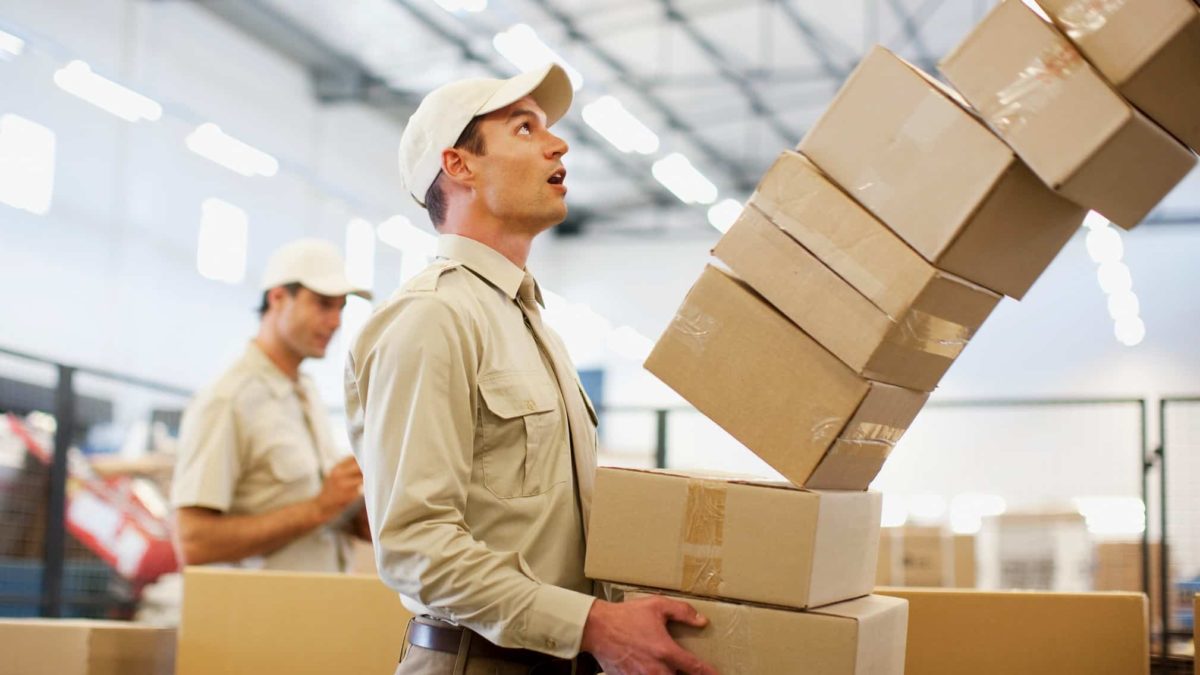 A man carrying a large stack of boxes watches with a wide-mouthed expression as the stack tumbles forward into the air.
