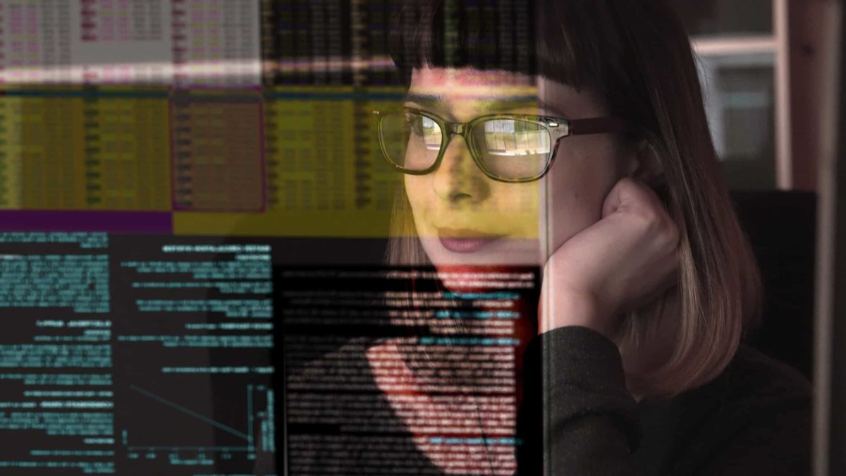 a woman with a small satisfied smile on her face looks at reflected, illuminated data on screens in front of her.