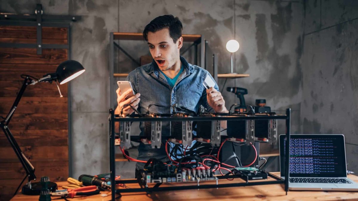 a cryptocurrency blockchain miner acts with surprise upon looking at his phone while standing behind a conglomeration of technology to access cryptocurrency.