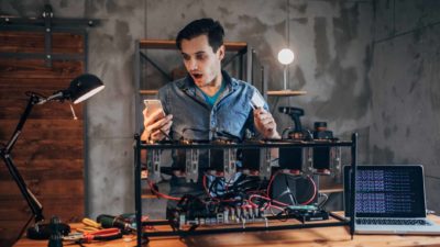 a cryptocurrency blockchain miner acts with surprise upon looking at his phone while standing behind a conglomeration of technology to access cryptocurrency.