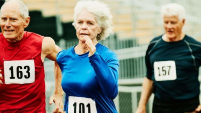 three older people wearing athletic outfits with racing numbers race around the bend of an athletics track.