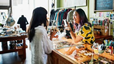 a woman produces her phone and shows it to the attendant at a shop counter as they appear to be in friendly conversation in a fashion boutique with clothes and accessories.