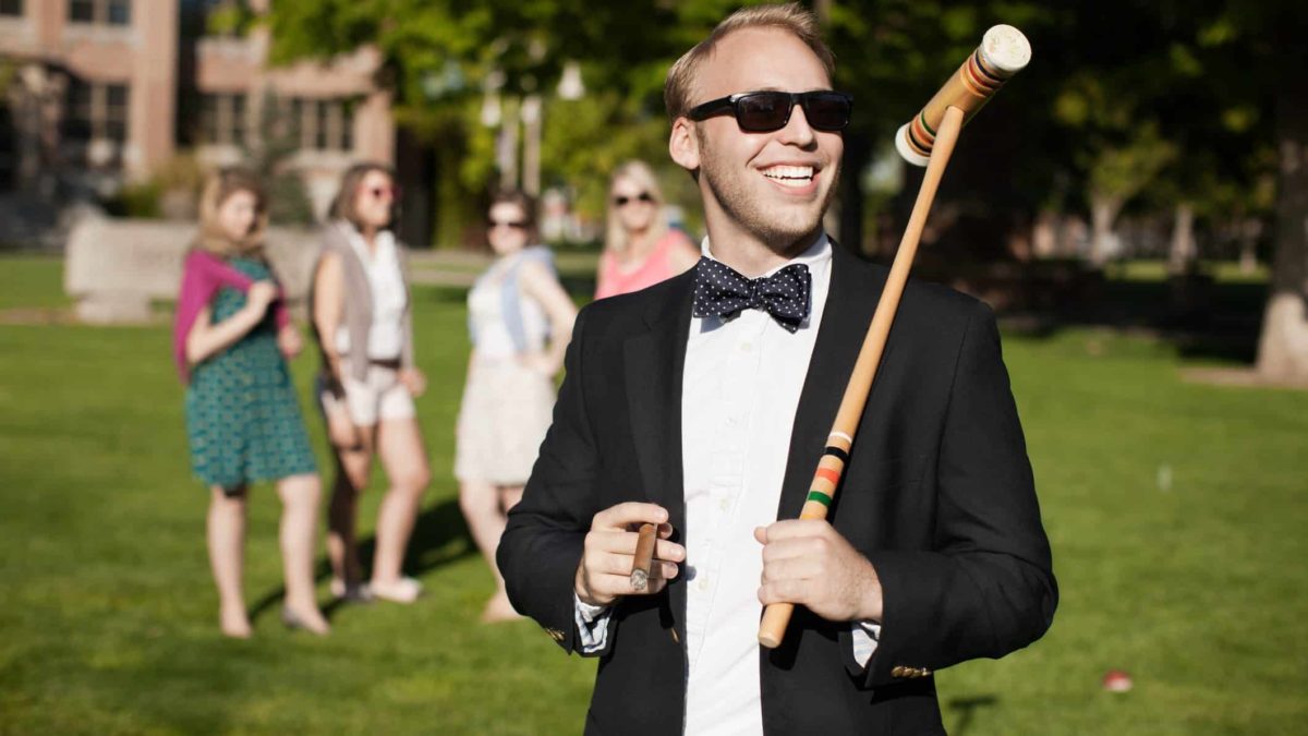 a man wearing a dinner suit holds a cigar and a croquet mallet on a rolling green lawn with a group of women and a mansion in the background.