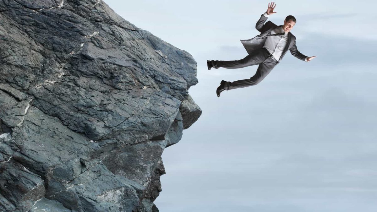 A man dressed in business suit freefalls from a rocky cliff with a grey sky background.