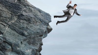 A man dressed in business suit freefalls from a rocky cliff with a grey sky background.
