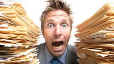 a man peers between two large piles of papers and files with a wide-eyed, wide-mouth look of dread at the amount of work he has to do.