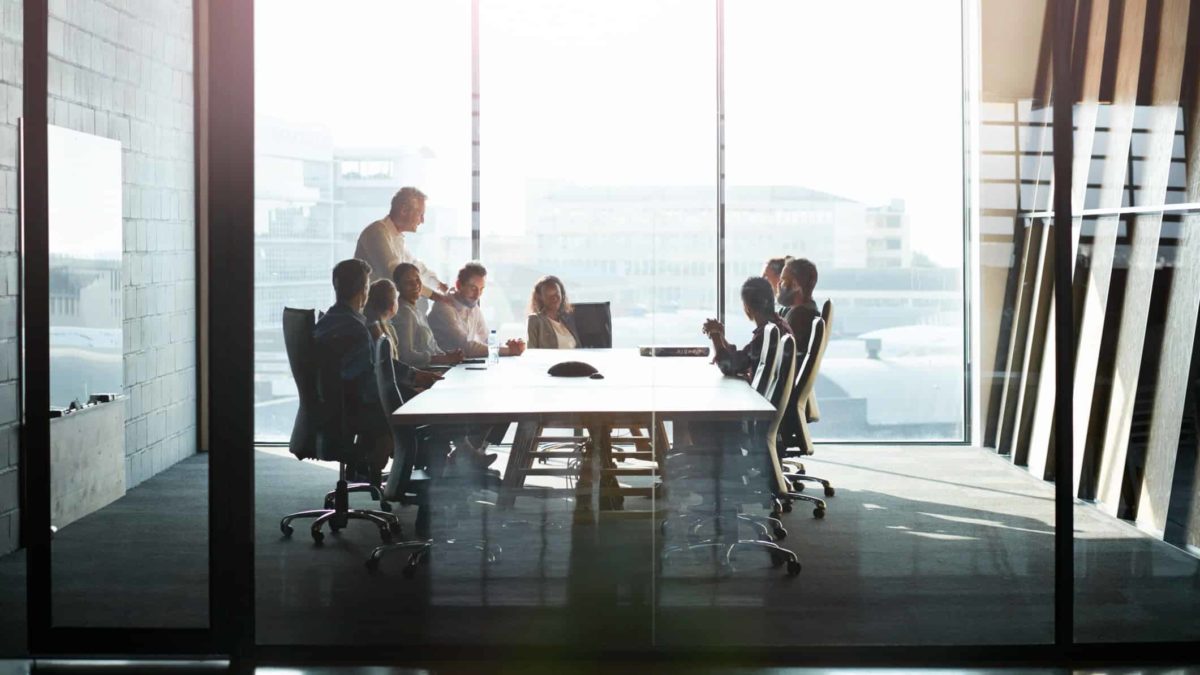 a board room with members sitting around a long table with one person standing and a large floor length window in the background showing a light-drenched cityscape view.