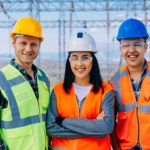 a group of three electricity workers stand smiling wearing hard hats and high visibility vests in front of an array of high voltage power equipment.