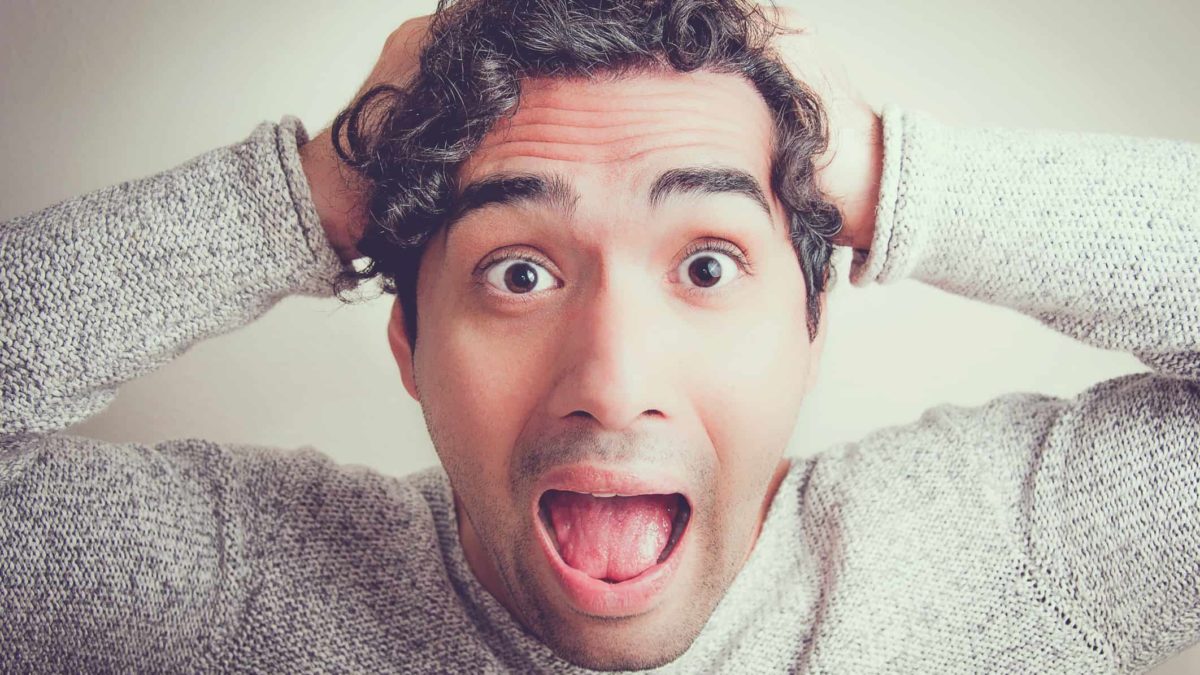 a close up of a man with wide open eyes and wide open mouth holding his head and reacting in shock and surprise to some share market ews.