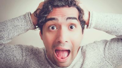 a close up of a man with wide open eyes and wide open mouth holding his head and reacting in shock and surprise to some share market ews.