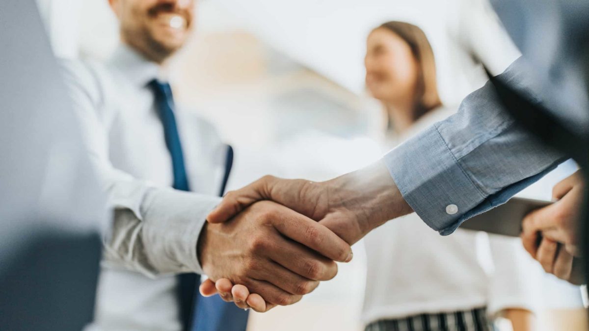 A close-up of a handshake depicting a business deal with one of the people in the background of the shot alongside a colleague looking pleased at the deal.