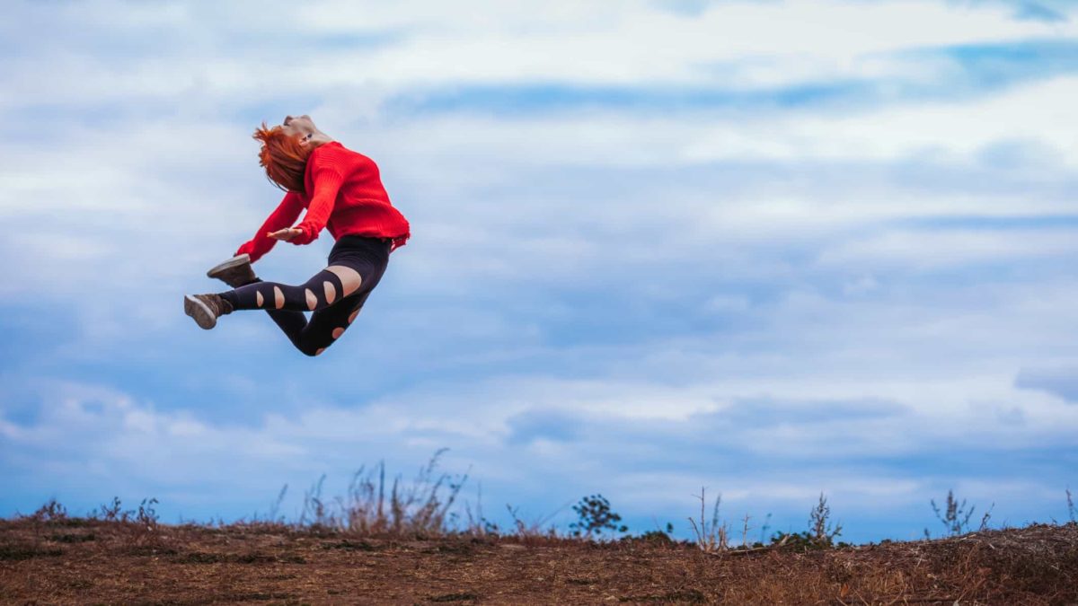 A woman wearing a red jumper leaps into the air with sky behind her and earth beneath her.