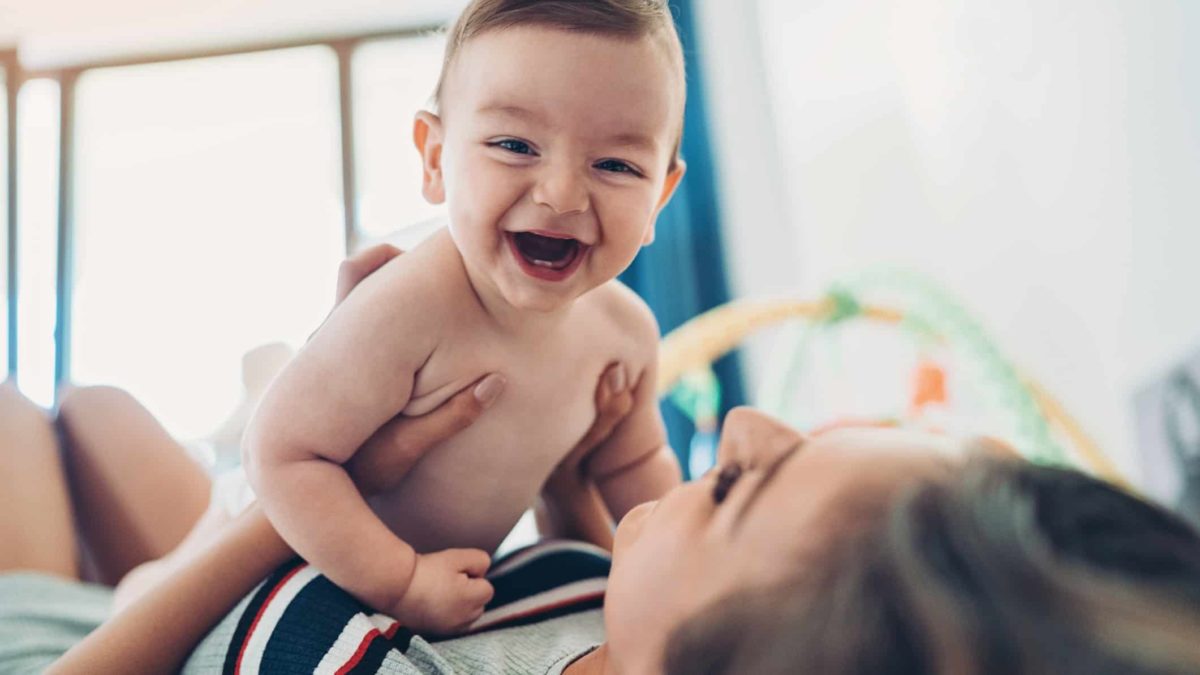 Mum playing with her baby boy holding him on her tummy as she lays down while smiling about the Bubs share price going up today