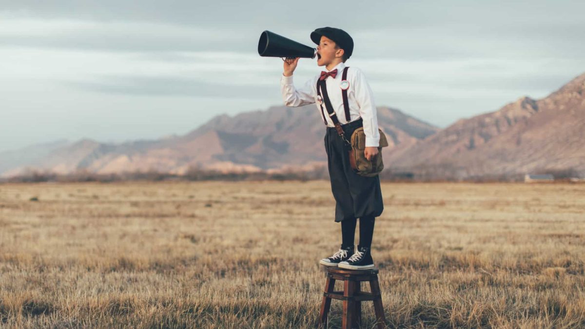 An old-fashioned news boy stands on a stool and yells through a microphone in an open field.