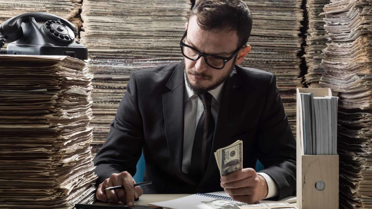 Adult man wearing a black suit and necktie calculating via old fashioned calculator, surrounded by newspapers.