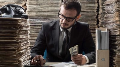 Adult man wearing a black suit and necktie calculating via old fashioned calculator, surrounded by newspapers.
